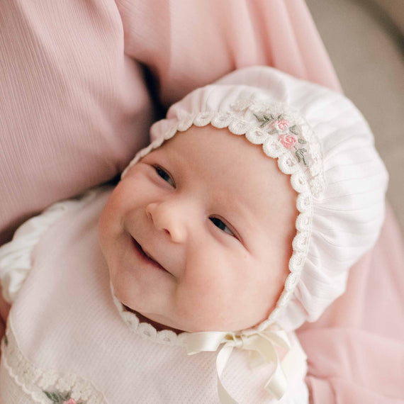 Baby girl wearing the Natalie Bonnet made with pink ribbed cotton with a cotton lace featuring embroidered pink rosettes and olive green leaves. A silk ribbon tie is strapped around her chin