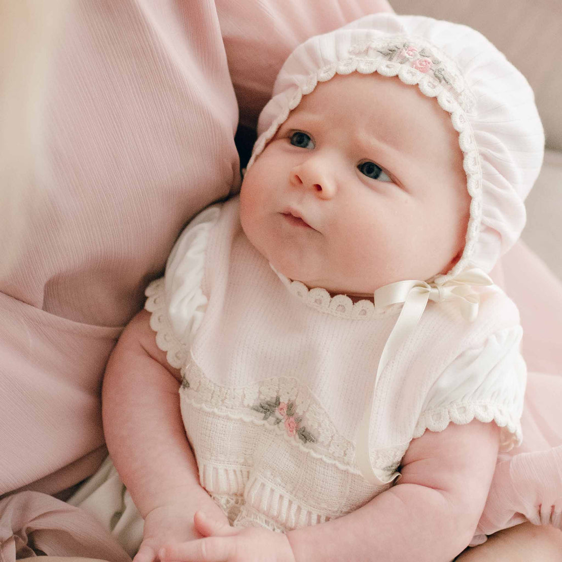 Baby girl wearing the Natalie Bonnet made with pink ribbed cotton with a cotton lace featuring embroidered pink rosettes and olive green leaves. A silk ribbon tie is strapped around her chin
