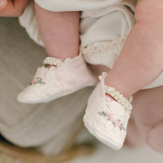 Baby girl wearing the Natalie Booties made from a pink textured cotton and a vintage natural cotton lace featuring embroidered pink rosettes and olive green leaf. A soft elastic band fits across her feet