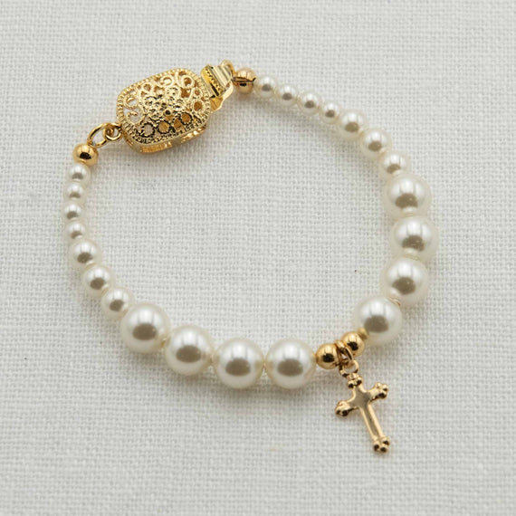 The Cream Luster Pearl Bracelet with Gold Cross features a gold filigree clasp and a gold-plated cross pendant, forming an elegant circle. Set against a white textured background, this delicate piece makes the perfect Christening gift.