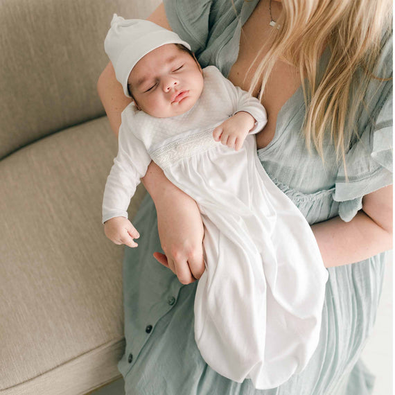 Photo of a newborn baby cradled in his mother's arms. He is wearing the Harrison Newborn Gown and Harrison Knot Cap. The Harrison Newborn Gown is made with soft pima cotton in white and features a plush white quilt bodice. Along the waist is an ivory Venice lace accent and features an open skirt design  