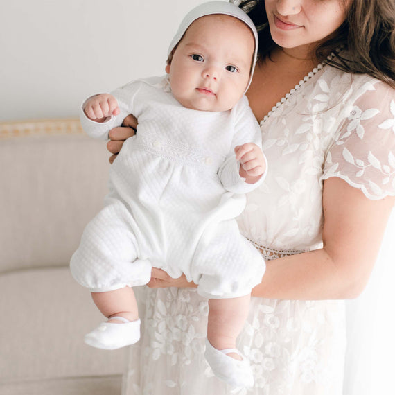 Mother holding her newborn baby who is wearing the Elijah Newborn Romper made from a plush white quilted cotton, featuring white Venice lace at the bodice and white linen trim at the cuffs and neck