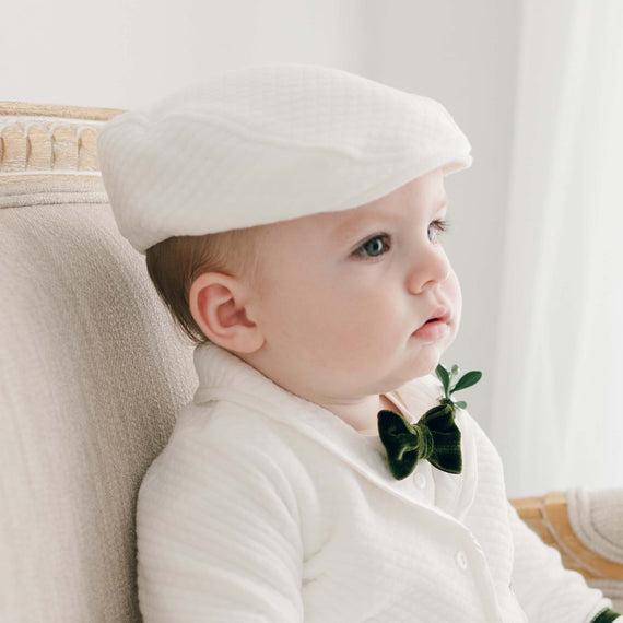 A profile view of a baby dressed in an heirloom, white textured sweater and a  Noah Ivory Newsboy Cap, holding a small green plant, sitting in an upscale cream-colored armchair.