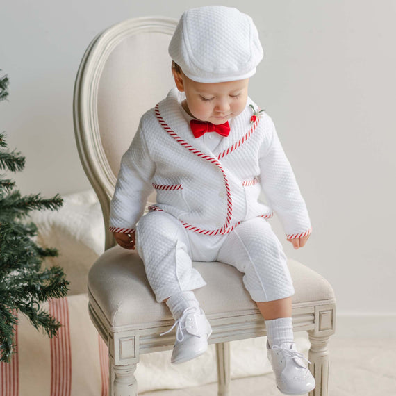 A baby wearing the Nicholas 3-Piece Suit, featuring red accents and a red bow tie, sits on a cream-colored chair. The child is also dressed in a matching white cap and shoes. This charming Christmas attire is beautifully complemented by the decorated tree partially visible in the background.