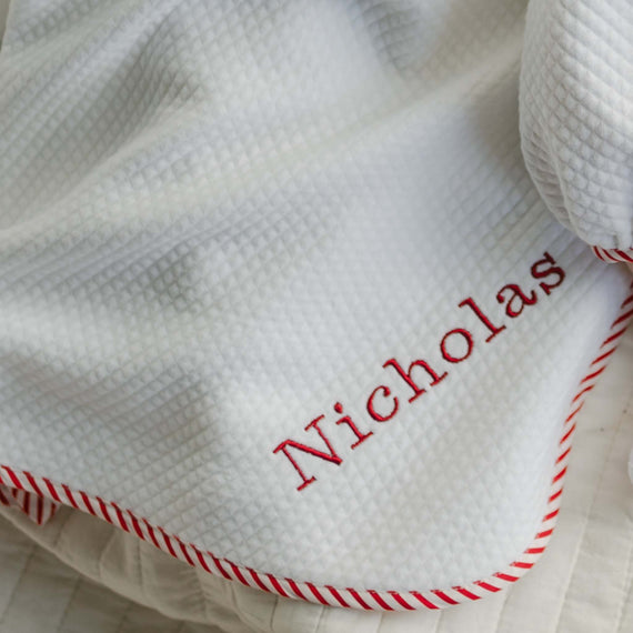 Introducing the Nicholas Personalized Blanket: a handmade white textured blanket featuring red striped edging and "Nicholas" embroidered in red thread. This unique blanket is partially folded, highlighting its soft texture.