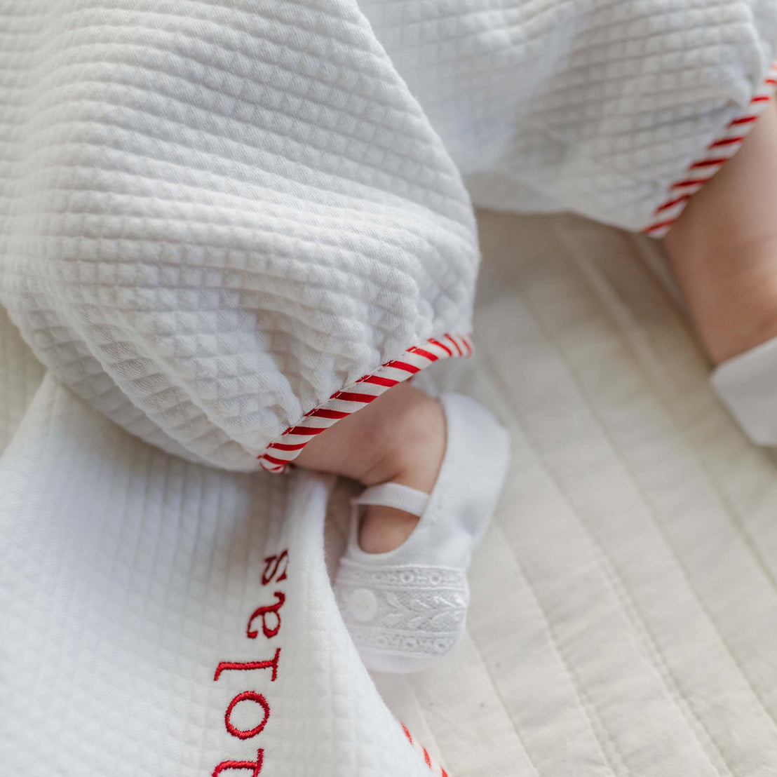 A baby in a white textured outfit featuring red trim and Venice lace lies on a quilted surface. The baby's name, partially visible, is embroidered in red on the fabric near their hand, which is wearing a Nicky Bootie.