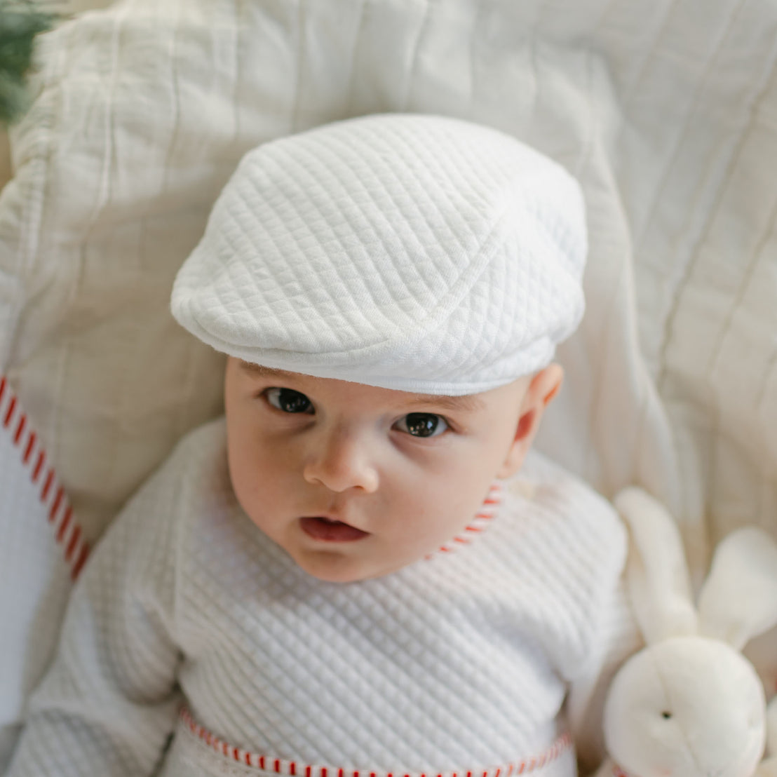 A baby wearing a quilted cotton outfit and the Nicholas Quilted Newsboy Cap is lying on a white blanket, looking up. A plush white rabbit toy is tucked next to the baby.