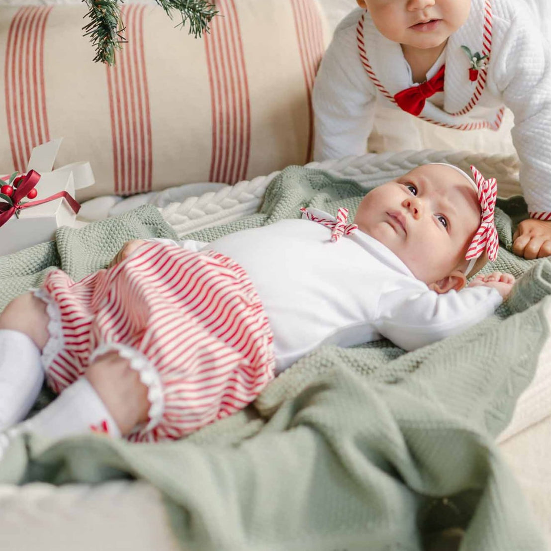 Two babies dressed in the Nicole Bloomer Set are near a decorated Christmas tree. One baby is lying on a pillow basket, while the other, in a red and white striped cotton onesie set and cap, sits nearby. The setting is cozy with a striped cushion and soft blanket.