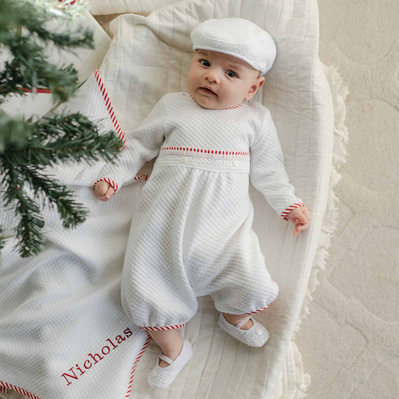 A baby wearing the Nicky Quilted Romper and matching hat lies on a cream-colored blanket with red trim, embroidered with the name "Nicholas." A Christmas tree branch appears in the foreground, perfectly capturing the essence of holiday season clothing.