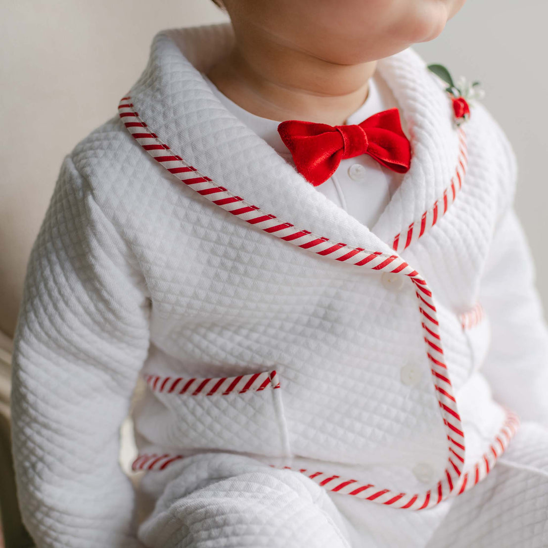 A young child is elegantly dressed in the Nicholas 3-Piece Suit, featuring a quilted cotton design with red trim and a charming red bow tie, making it perfect for Christmas. This suit, which could be handmade, includes red accents on the collar and pockets and is adorned with a small decorative plant pin on the lapel. 