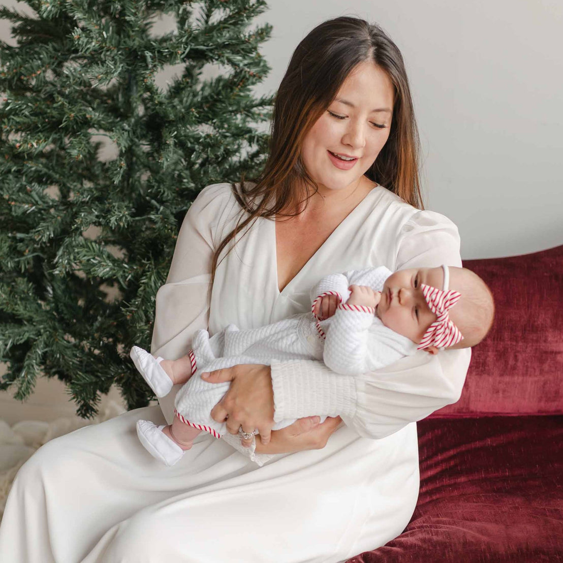 A woman in a white dress cradles a sleeping baby dressed in the Nicky Quilted Romper, featuring red and white striped accents. They are seated on a burgundy cushion next to a partially decorated Christmas tree, embodying the warmth of the holiday season.