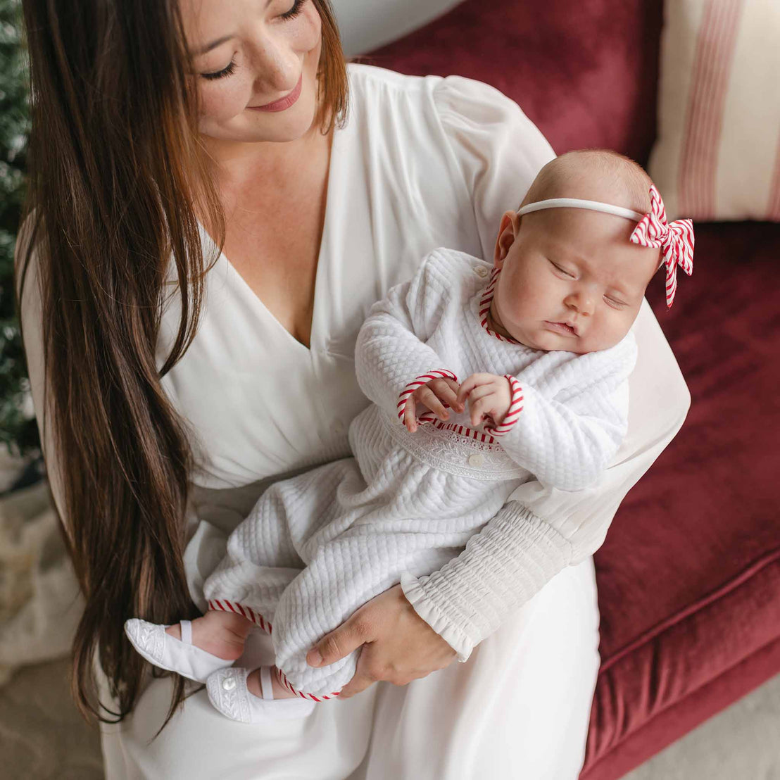 A woman with long brown hair, dressed in a white gown, cradles a sleeping baby wearing the Nicky Quilted Romper, featuring red striped accents. The matching headband completes their festive look as they sit on a plush burgundy couch, enhancing the cozy ambiance.