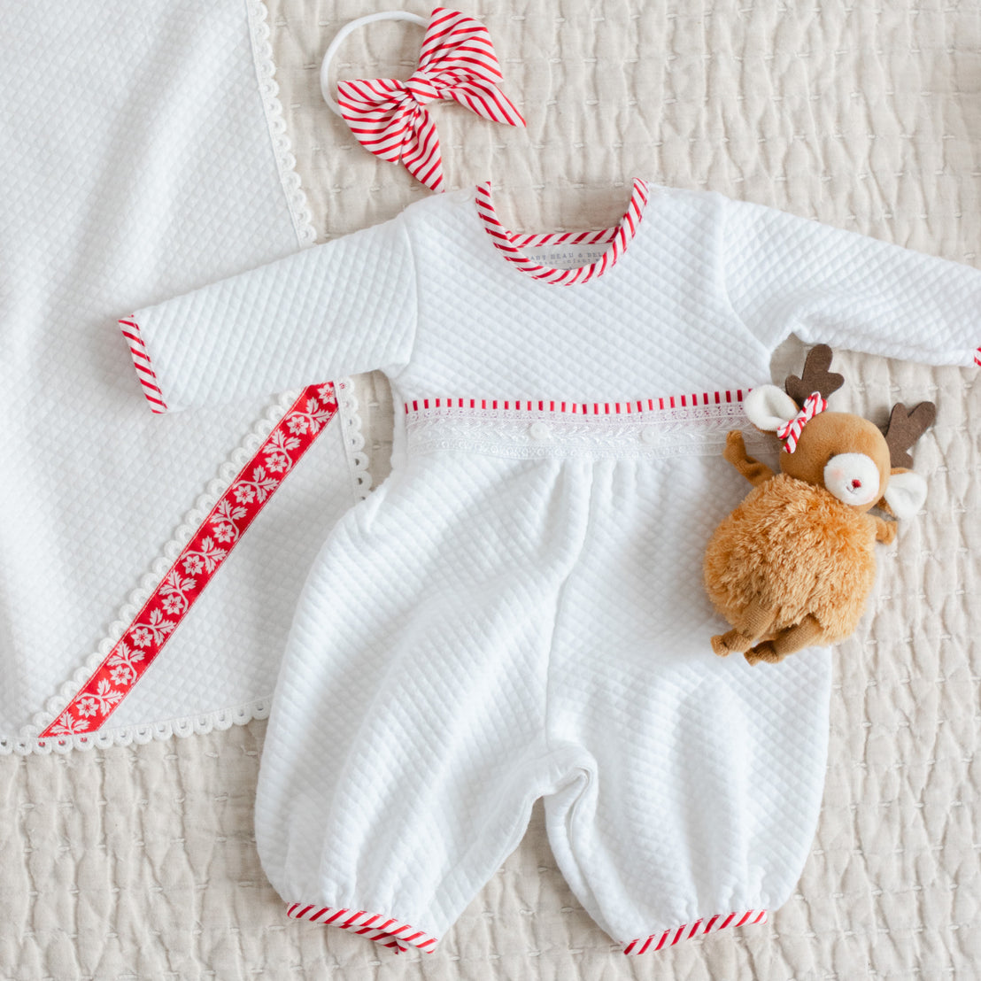 White baby romper with red trim and matching headband bow alongside a plush reindeer toy and white quilt blanket.