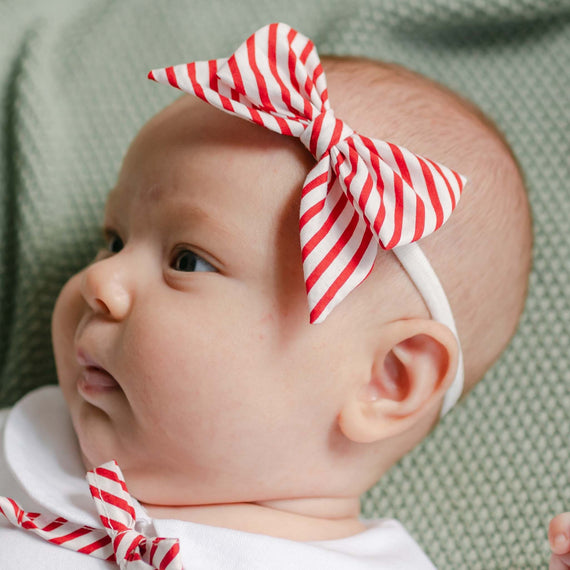 A baby wearing a Nicole Bow Headband, featuring a red and white handmade design, lies on a green textured surface in a white outfit. The striped cotton pattern adds an adorable touch as the baby gazes to the side.