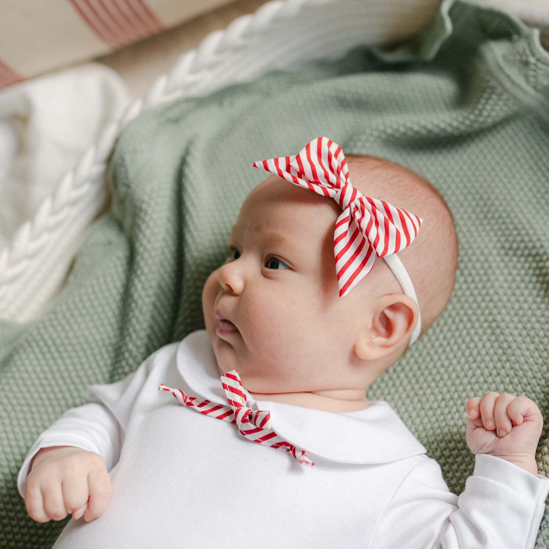 A baby wearing a white outfit and a handmade Nicole Bow Headband in red-and-white striped cotton lies on a green textured blanket, looking to the side with a curious expression.