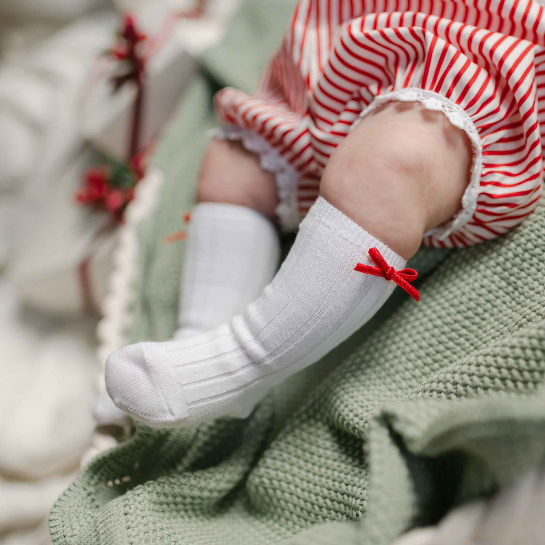 Dressed in a charming Christmas outfit, a baby relaxes on a soft green blanket, wearing red and white striped leggings and Nicole Knee Socks embellished with red velvet ribbons.