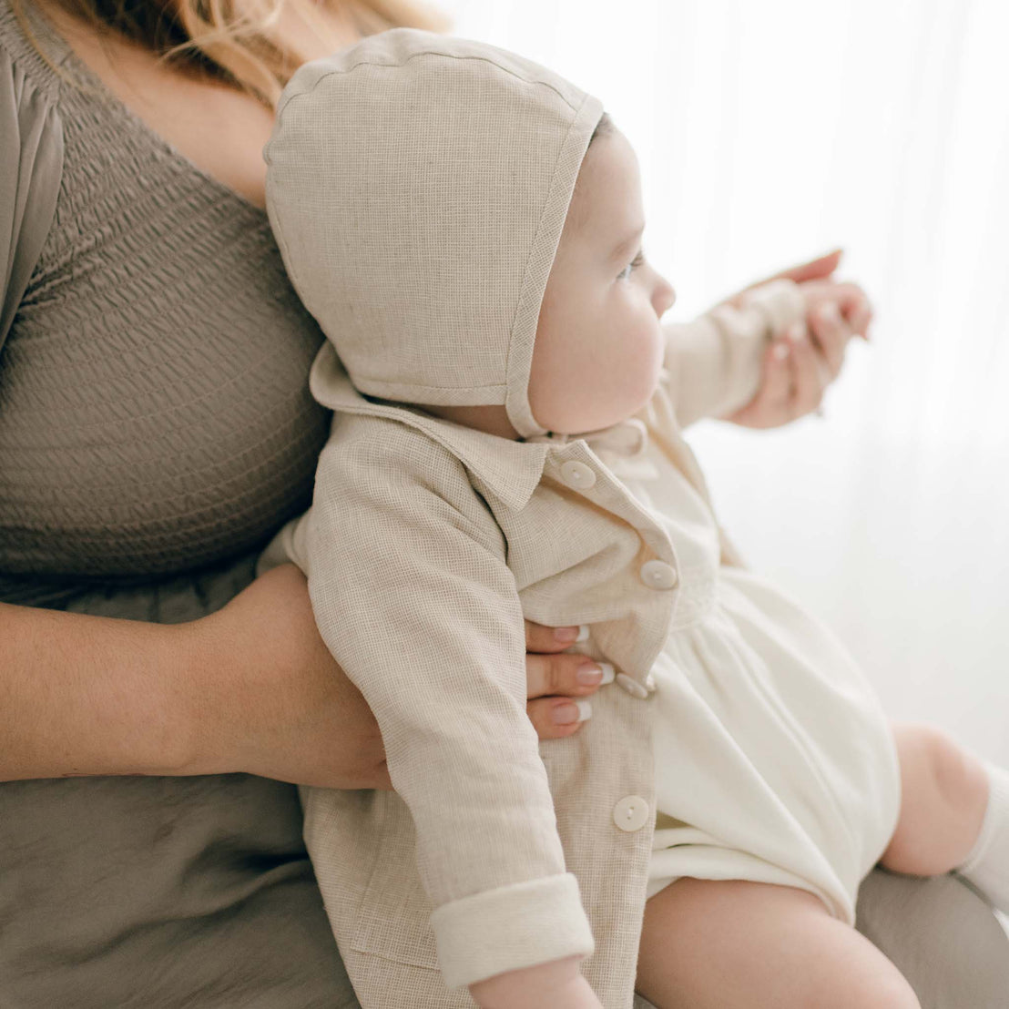 A baby dressed in a beige French terry cotton jacket and bonnet sits on a woman's lap. The woman is wearing the Henry Trench Coat, featuring a textured gray design. The background is softly lit.