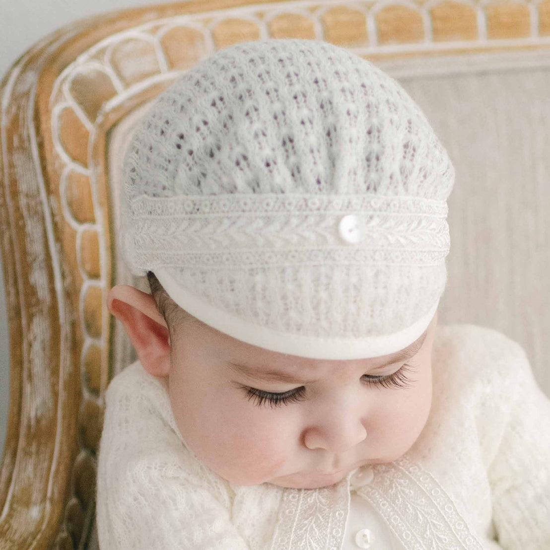 A baby dressed in the Oliver Romper and Knit Sweater and Cap ensemble in ivory featuring intricate embroidery. He is sitting on a light-colored upholstered armchair. The baby is looking down away from the camera.