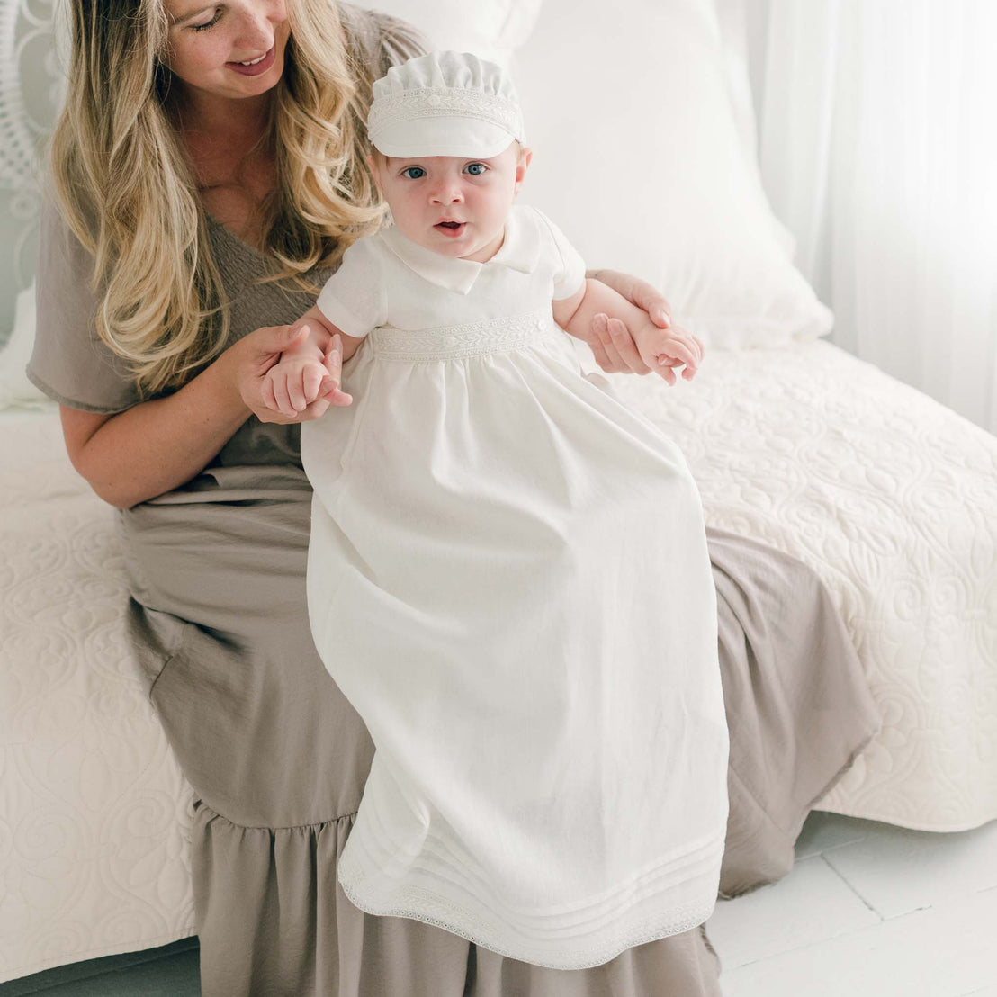 A woman with long blonde hair, wearing a light gray dress, sits on a white bed holding the hands of a baby dressed in an Oliver Convertible Skirt adorned with Venice lace. The baby looks at the camera with a curious expression. The setting appears bright and serene.