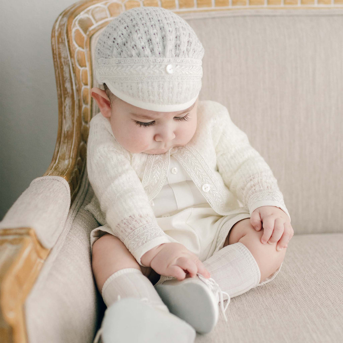 A baby in the light ivory Oliver Sweater Shorts Suit sits on a beige vintage sofa, looking down at their Boys Ivory Two Tone Wingtip Shoes.