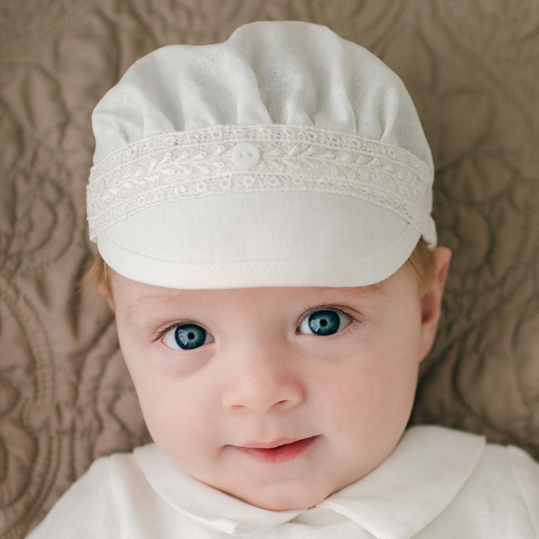 Close-up of a baby with big blue eyes, wearing an Oliver Linen Cap with exquisite Venice lace detailing and a button in the center. The baby is dressed in a white outfit, lying on a textured beige surface. The baby's expression is calm and slightly smiling.