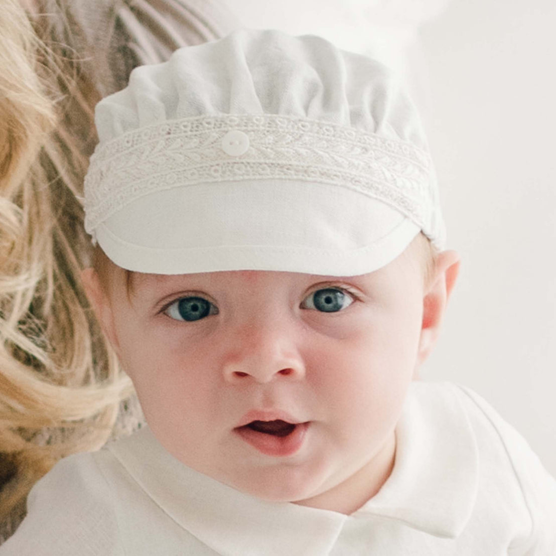 A baby wearing an Oliver Linen Cap, handmade in the USA, and a matching white shirt looks directly at the camera with blue eyes and a slight open-mouthed expression. Blond hair of an adult is partially visible on the left side of the image.