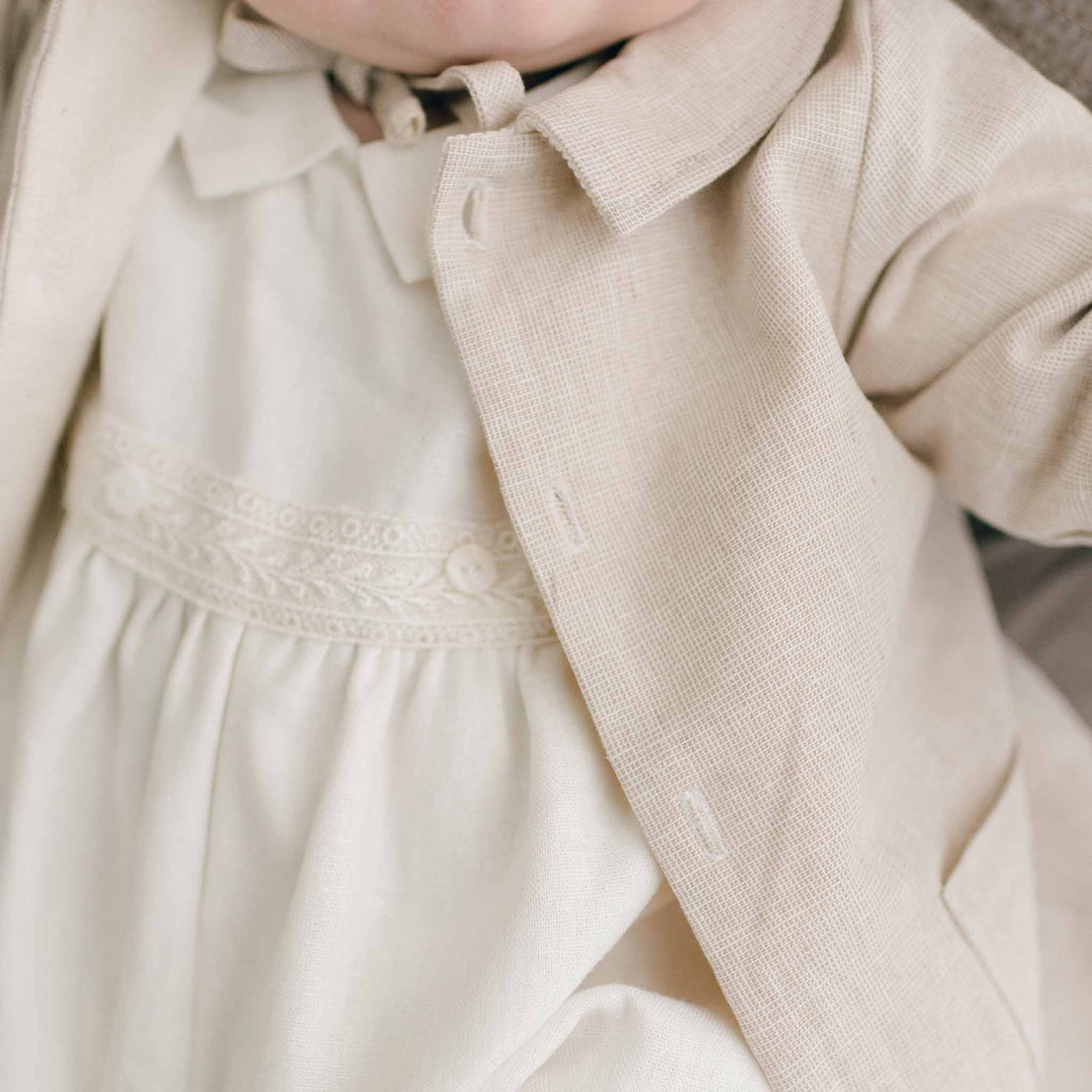 Close-up image of a baby wearing the Henry Trench Coat, a beige linen piece, over a cream-colored outfit with delicate lace detailing. This handmade garment from the USA looks soft and formal, capturing a classic and elegant style.