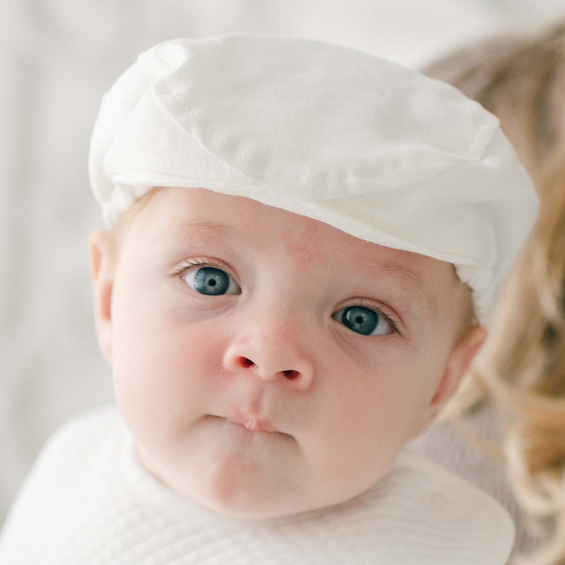 A close-up of a baby with blue eyes wearing a handmade white christening outfit and an Oliver Linen Newsboy Cap. The baby has a serious expression and is looking directly at the camera. The background is soft and blurry, with a hint of an adult's shoulder visible.