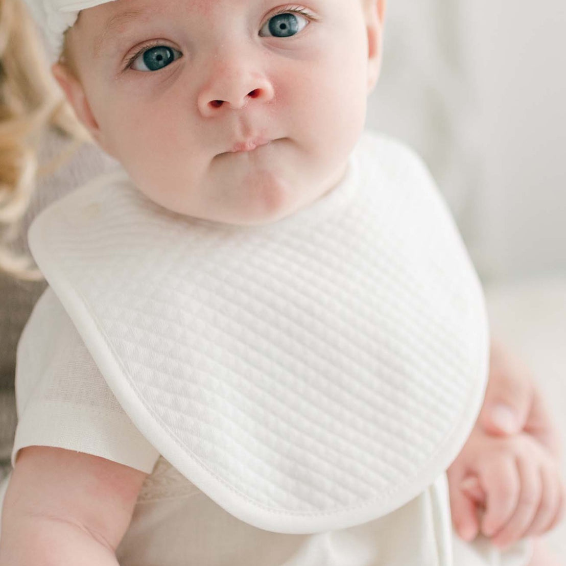 A baby with blue eyes wearing a white cap and an Oliver Bib is sitting on a woman's lap. The woman's face is not visible, and she has blonde, wavy hair. The baby looks directly at the camera with a slightly pouting expression.