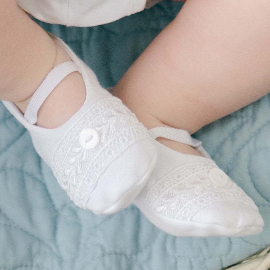 Close-up of an infant's feet wearing delicate, white, embroidered booties with buttons from the Oliver Knit Accessory Bundle - Save 15%. The baby is seated on a personalized quilted blanket. The intricate design on the booties showcases fine craftsmanship, adding a touch of elegance to the cozy scene.