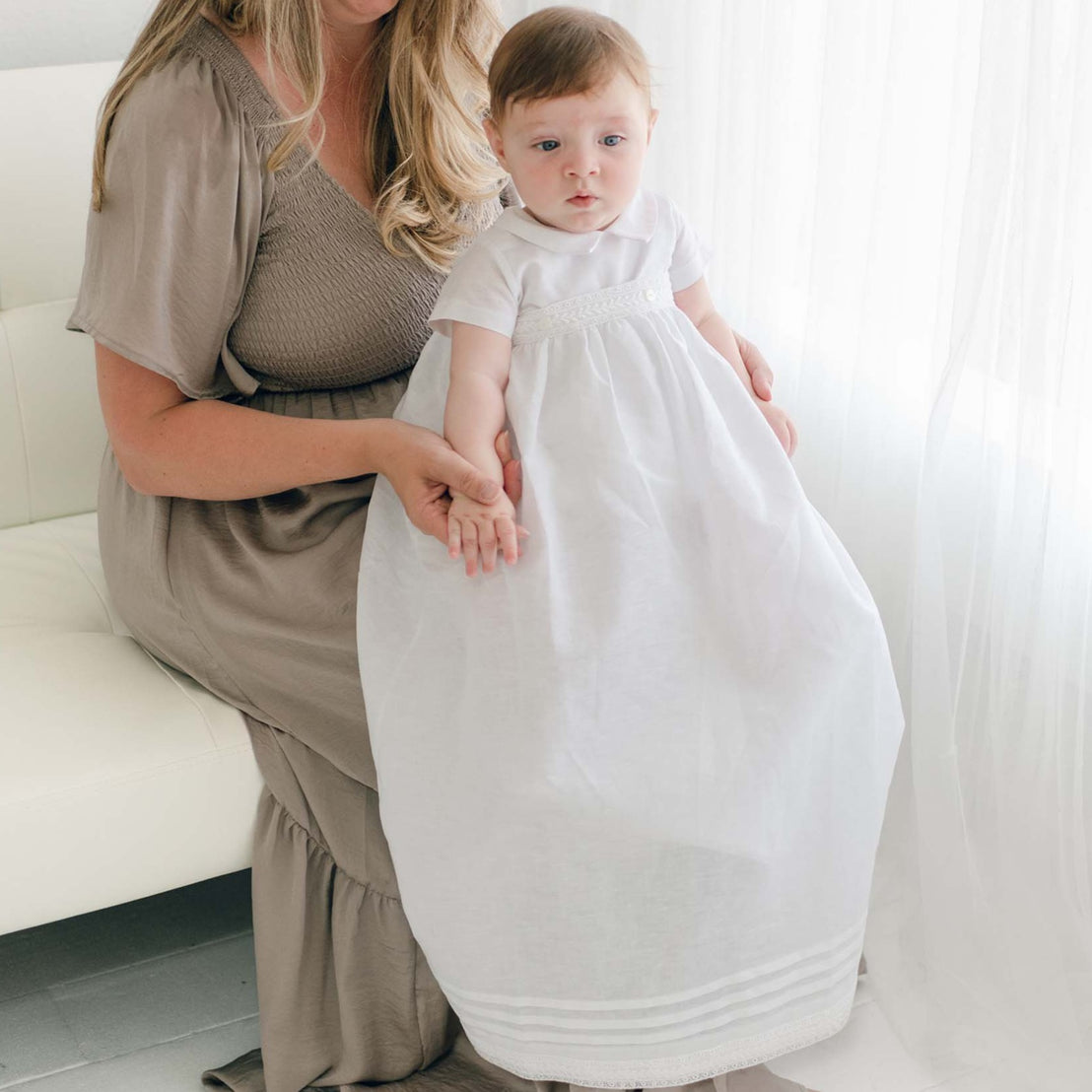 A woman with long blonde hair wearing a brown dress sits on a white couch, holding a baby dressed in an Oliver Convertible Skirt adorned with delicate Venice lace. The baby has light hair and looks forward while the woman smiles down at the baby. A sheer white curtain hangs in the background.