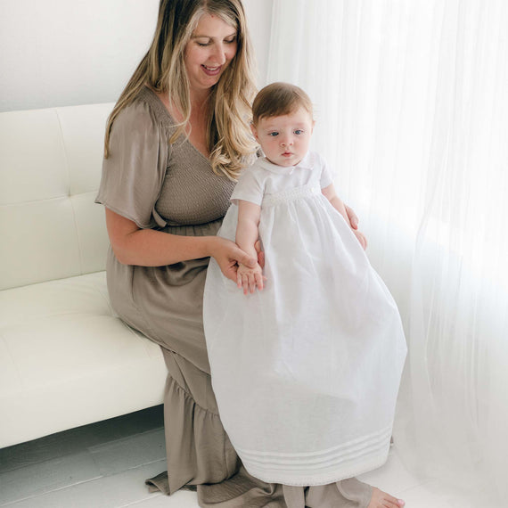 A woman with long hair, wearing a taupe dress, is sitting on a white couch. She is gently holding a baby dressed in an Oliver Convertible Gown | Romper & Skirt Set, who is standing on the floor. Natural light streams through a sheer curtain in the background.