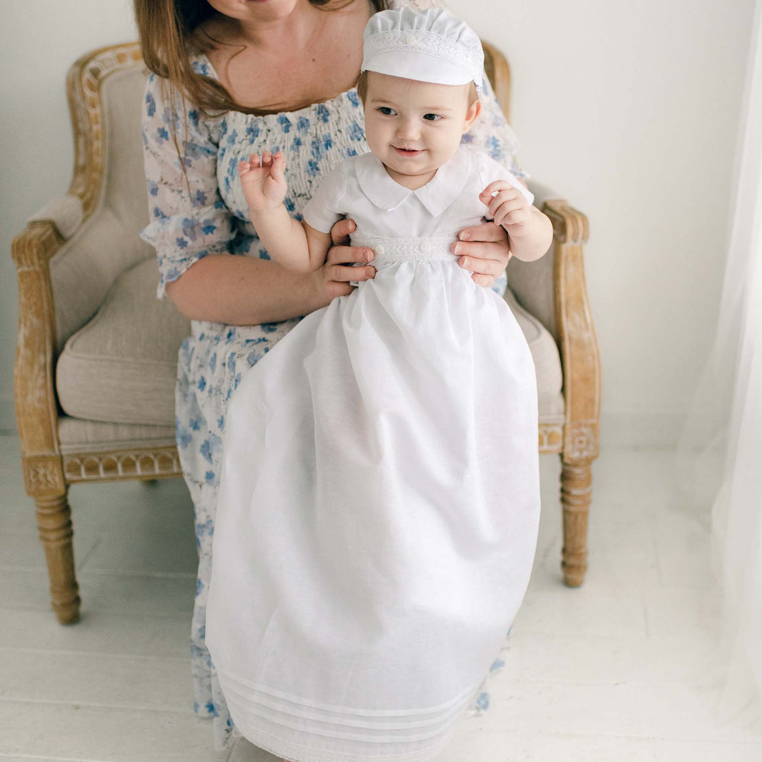 Baby boy sitting on a chair and wearing the White Oliver Convertible Gown and Romper (with matching Linen Newsboy Hat).