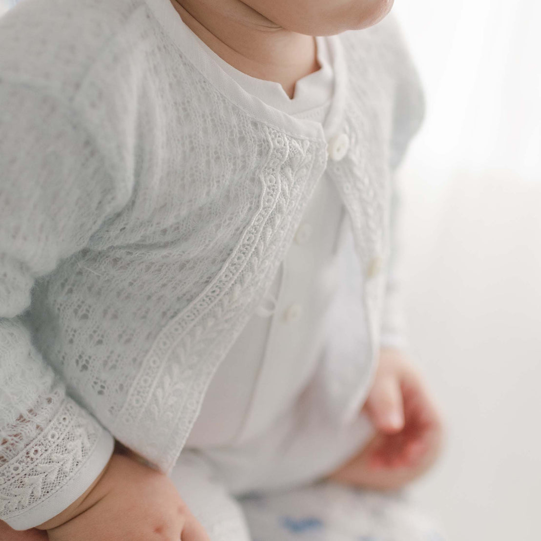 Close-up of a baby boy wearing a light blue knit Oliver Sweater, focusing on the knit sweater and matching lace and linen trim. The background is softly blurred.