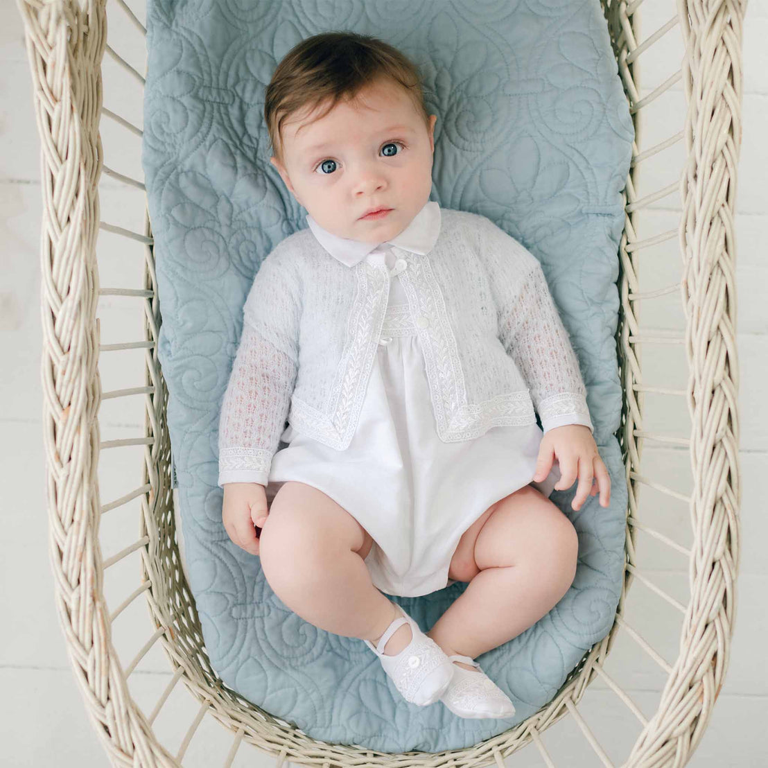 A baby with blue eyes and sandy brown hair wearing a white outfit and an Oliver Knit Sweater is lying down in a wicker basket on top of a quilted light blue blanket, looking up at the camera. One of the baby's legs is slightly bent.