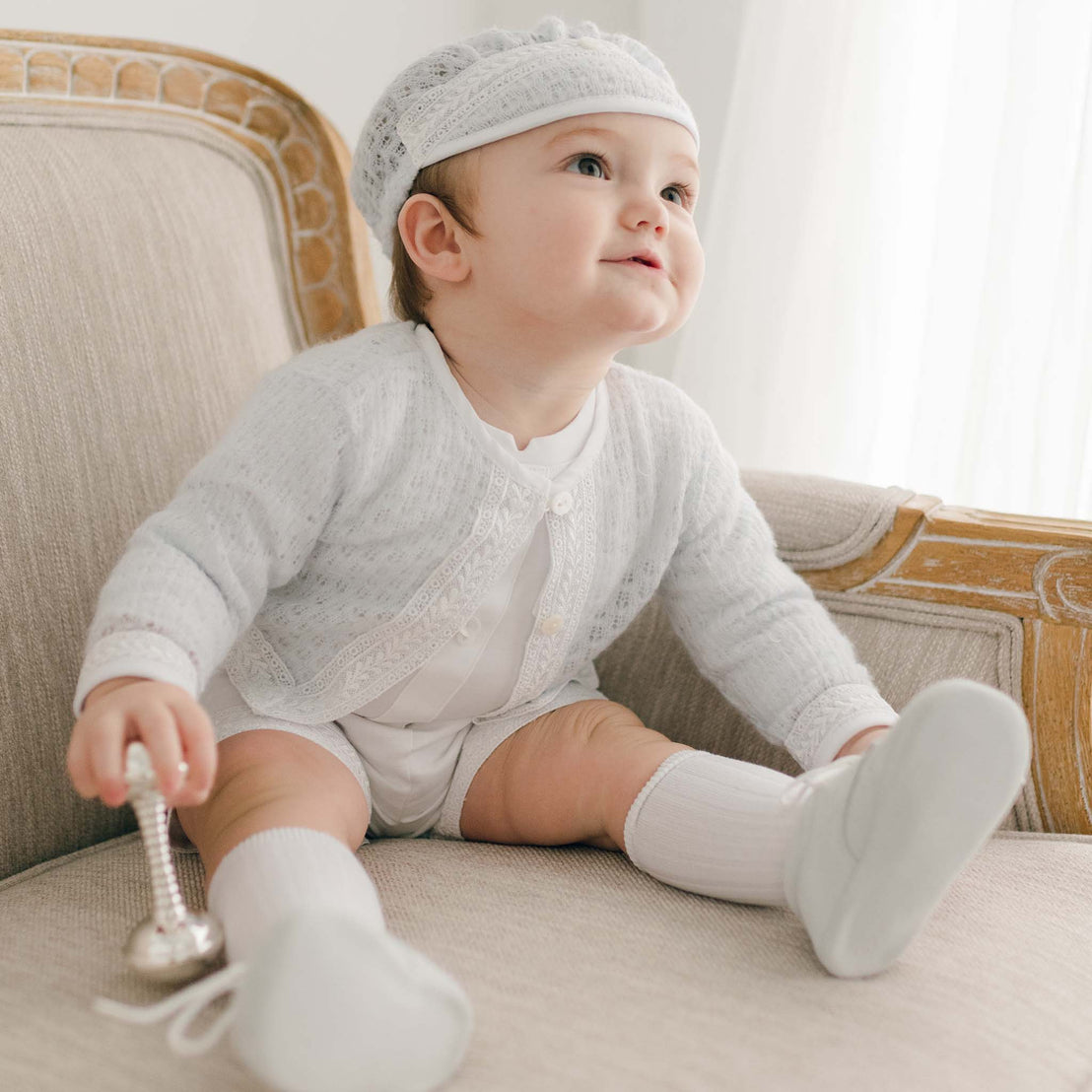 A joyful baby boy wearing the blue knit Oliver Sweater Shorts Suit sits on a beige sofa, holding a silver rattle, and looking up with a bright smile.