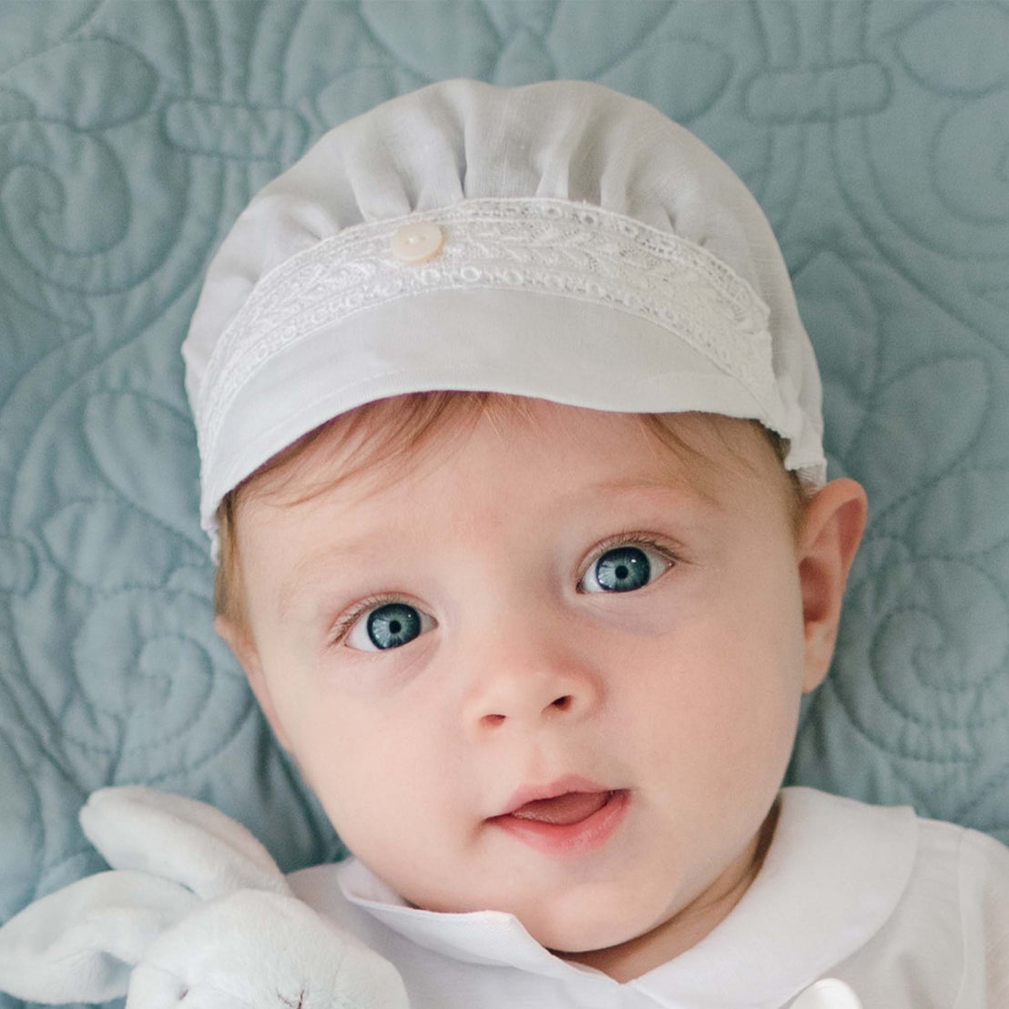 A baby with bright blue eyes wearing a handmade Oliver Linen Cap and a white outfit lies on a soft, light blue patterned blanket. The baby gazes directly at the camera with a small plush toy visible at the bottom left of the image.