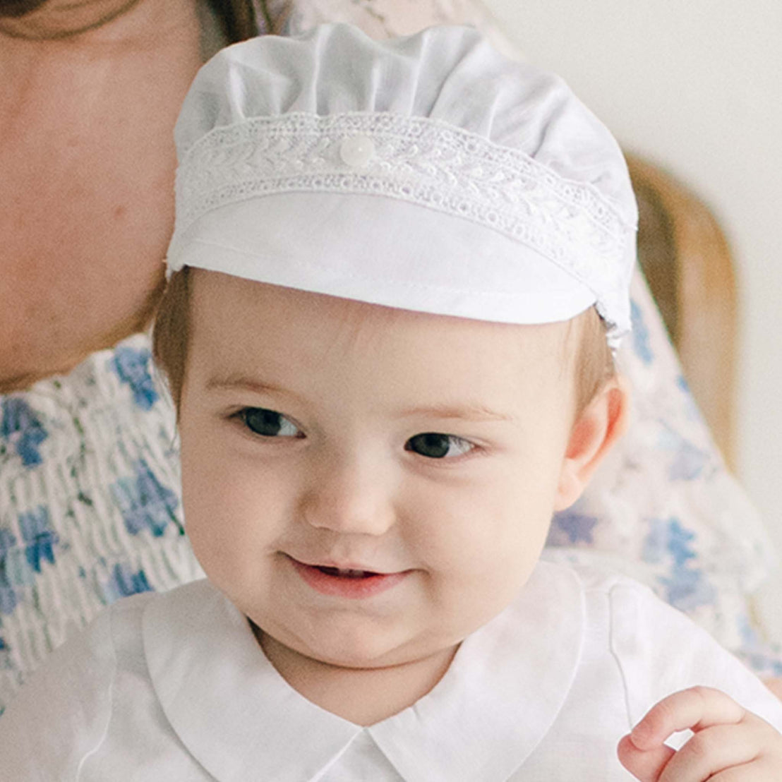 A smiling baby wearing an Oliver Linen Cap and a matching outfit with Venice lace details shines brilliantly. The baby is cradled by someone dressed in a floral-patterned blouse, adding to the heartwarming scene.