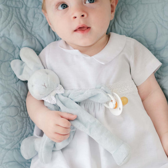 A baby wearing the Oliver Romper and matching linen cap in white. He is lying on a light blue quilted blanket, holding a blue Oliver Silly Bunny Buddy pacifier holder. The silly bunny is wearing a matching Oliver Lace Bow Tie.