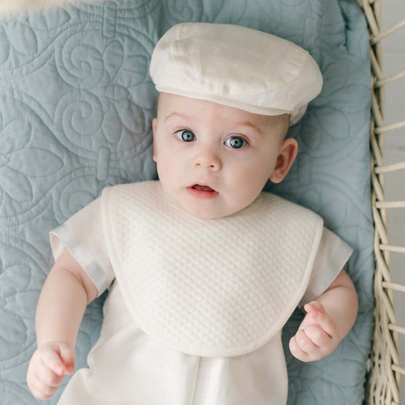 A baby boy wearing the Owen Bib over the Owen Linen Romper lies on a teal quilted cotton blanket. The baby's blue eyes look up with a curious expression.