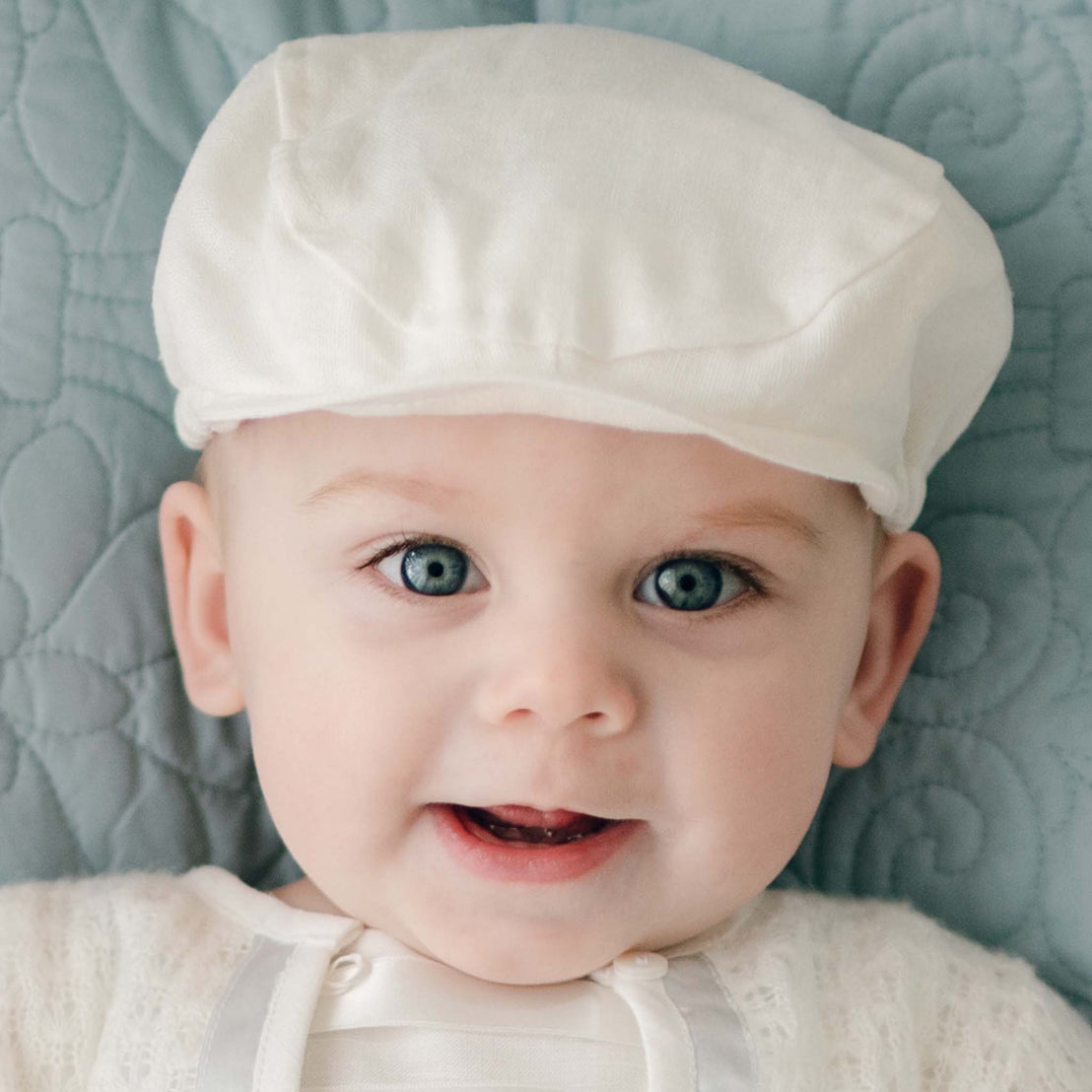 Close-up of a baby with bright blue eyes, smiling while wearing an Oliver Linen Newsboy Cap and matching white outfit. The background is a light blue quilted fabric.