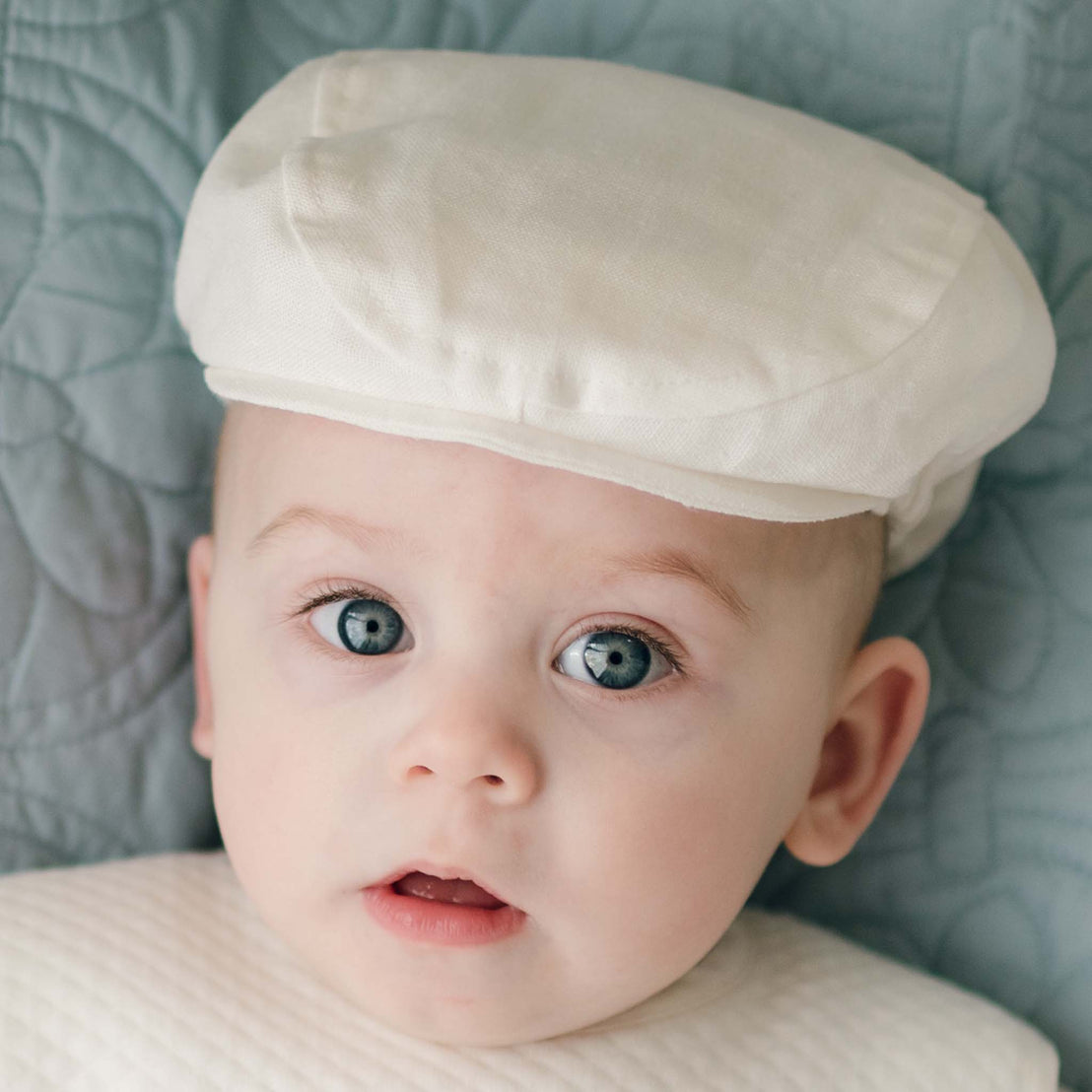 A baby with big blue eyes is wearing a handmade white christening outfit and looking at the camera. The baby, topped with an adorable Oliver Linen Newsboy Cap, is laying against a light gray quilted background.