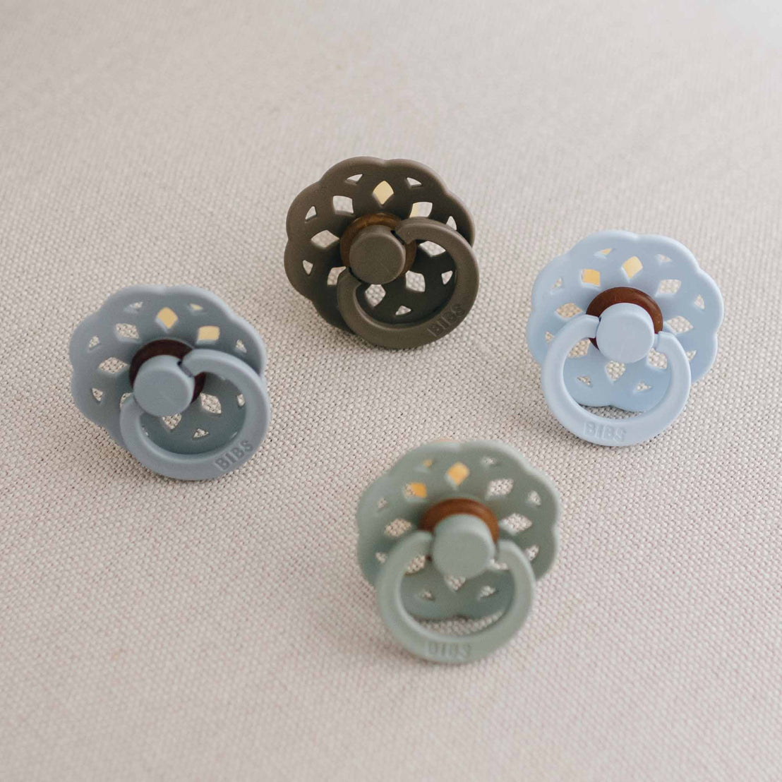 Five Bibs Lace Pacifiers, each with bohemian inspired designs and colors, arranged in a circle on a light beige fabric.