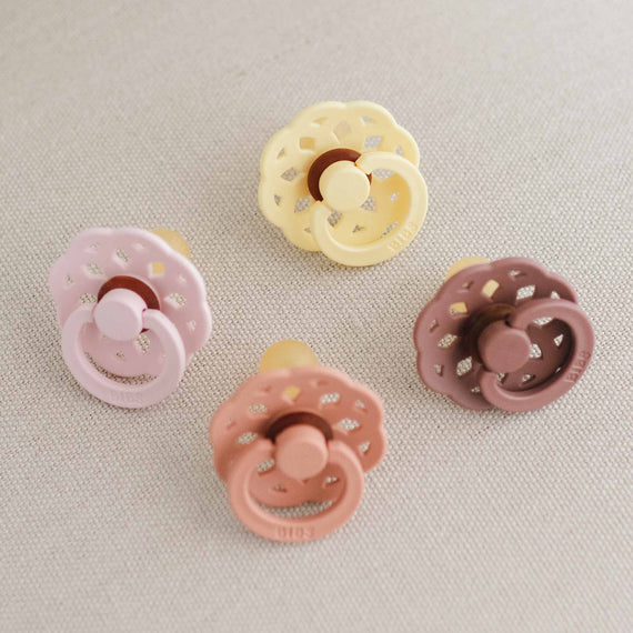 Four Bibs Lace Pacifiers in soft colors of blossom, peach, and sunshine woodchuck, each with a different design, neatly arranged on a textured light beige background.