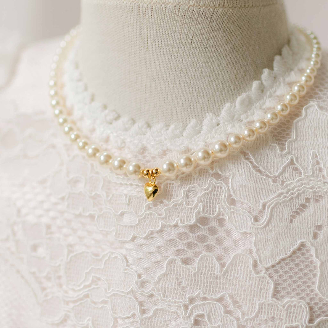 Pearl necklace with a gold heart on a mannequin wearing an ivory lace garment.