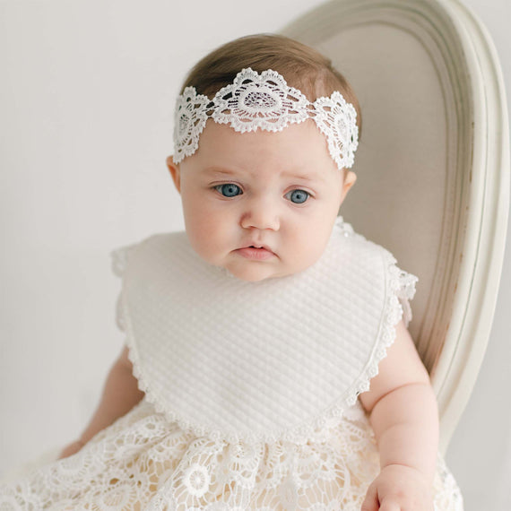 A baby with blue eyes sits on a cream-colored chair, wearing a white lace headband and a white lace dress with a quilted, handmade Poppy Bib. The ensemble offers the perfect baptism outfit, enhanced by its vintage touch. The background is softly blurred.