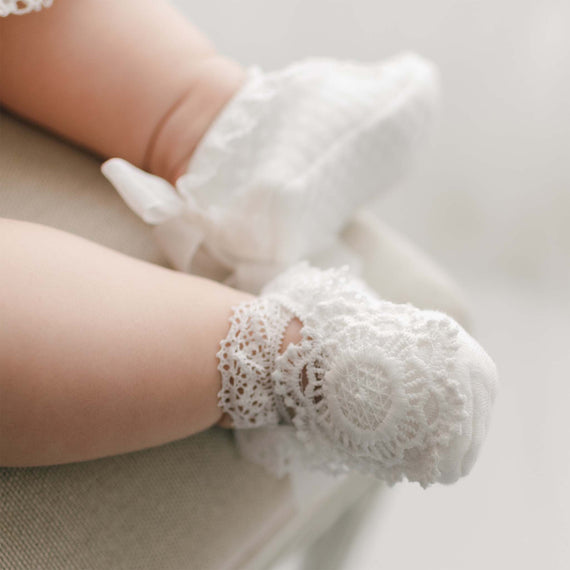 Close-up of a baby's legs wearing white, lace-trimmed Poppy Booties with floral lace applique. The baby is seated, and the background is softly blurred, focusing attention on the delicate details of the Poppy Booties and the baby's chubby legs.