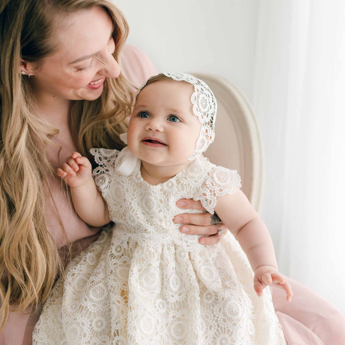 A woman with long blonde hair smiles and holds a baby dressed in a delicate white lace Poppy Christening Gown & Bonnet. The baby has blue eyes and looks up with a curious expression. The background is softly lit, creating a warm, intimate scene.