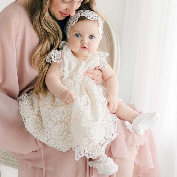 A woman in a light pink dress is holding a baby dressed in a Poppy Dress & Bonnet. They are sitting on a white chair next to a bright window. The baby, wearing white shoes with bows, looks directly at the camera while the woman gazes down affectionately.