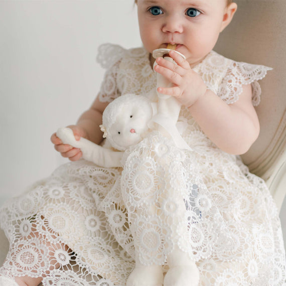 A baby girl with blue eyes, sitting on a chair, wearing a white lace dress. She is holding a Poppy Silly Lamb Buddy | Pacifier Holder while putting a golden pacifier in her mouth. The background is soft and neutral.